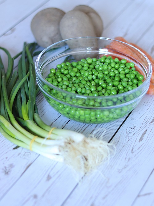 A medium-sized glass bowl full of frozen green peas ready to be prepared for lunch with green onions, carrots, potatoes and dry paprika.