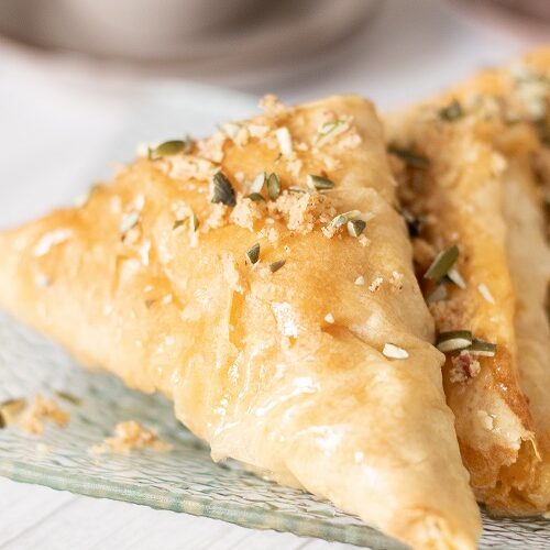 This blog post is about how to make baklava at home. Close up of piece of baklava garnished with chopped pumpkin seeds and nuts.