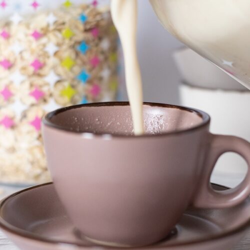 Pouring oat milk in a cup with a jar full of rolled oats in the background. Easy recipe of how to make oat milk.