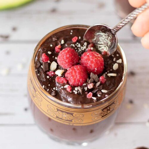 Vegan chocolate avocado pudding. Healthy and cream mousse topped with raspberries and chocolate flakes with a spoon.