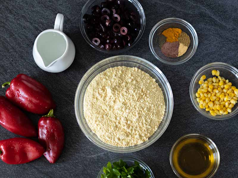 Plant-based-simple ingredients for baking no-yeast savoury muffins in tins.  Olives, chickpea flour, olive oil, water, red peppers, fresh parsley, corn and spices.