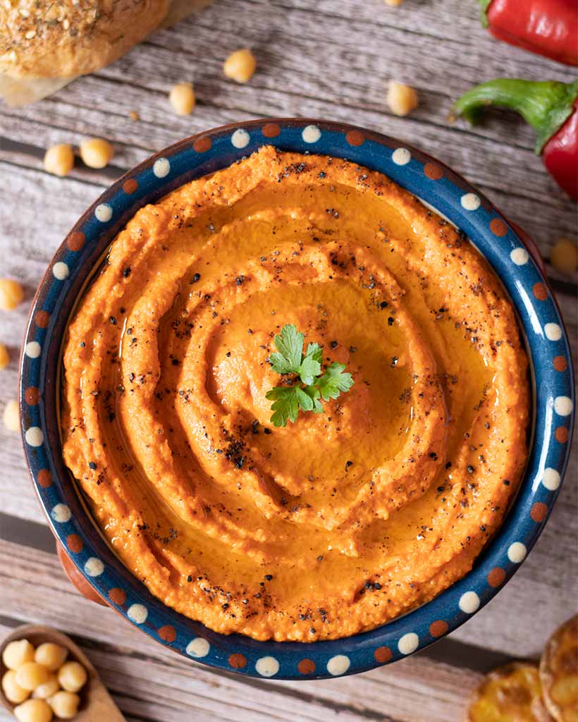 A healthy vegan dish full of colorful orange-red hummus made of roasted red peppers and chickpeas decorated with fresh parsley leaves and ground black seed on wooden table with a bagel and potato chips.
