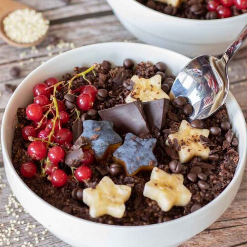 Vegan chocolate quinoa breakfast bowl with spoon topped with red currants, plums, bananas and dark chocolate chips on wooden table.