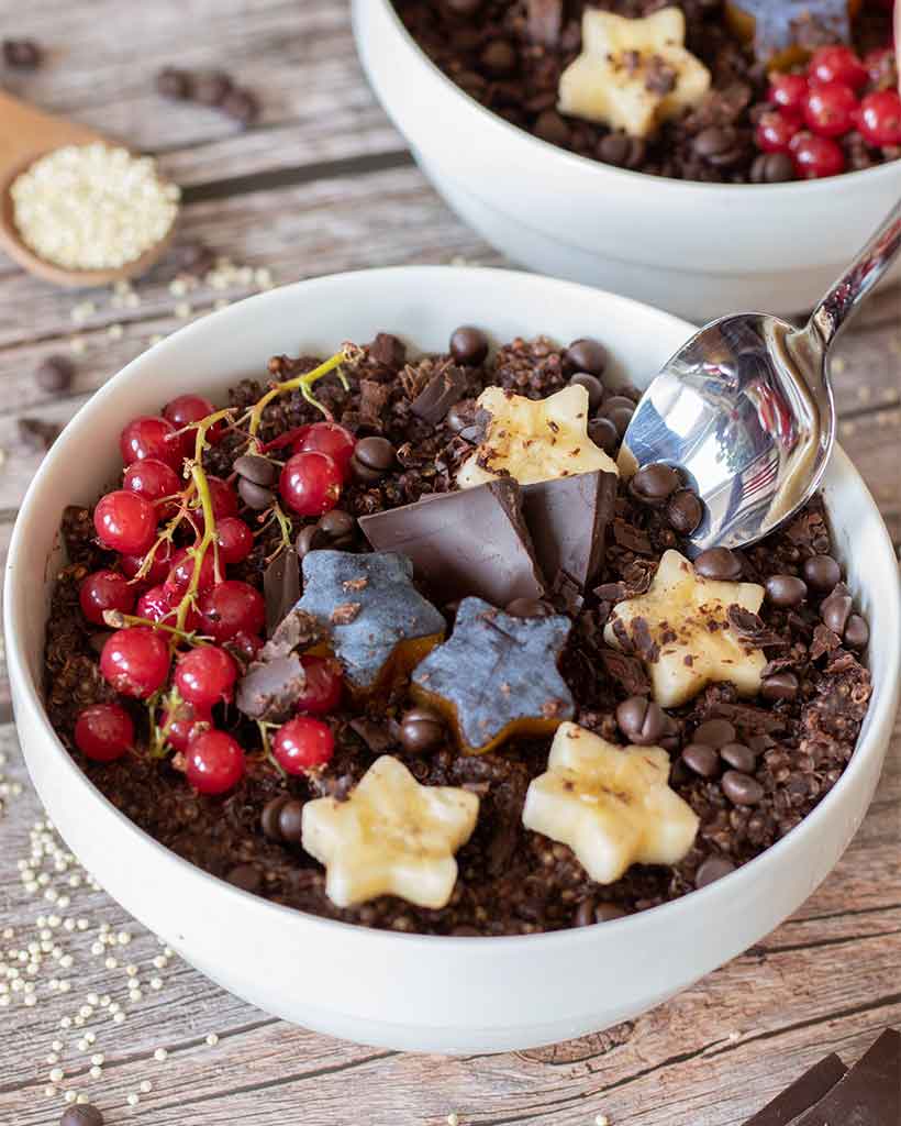 Vegan chocolate quinoa breakfast bowl with spoon topped with red currants, plums, bananas and dark chocolate chips on wooden table.