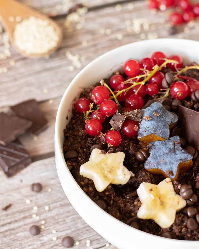 A close up of healthy chocolate quinoa porridge decorated with vegan dark chocolate squares and chips, banana, plums and red currants. Easy kid-friendly breakfast or dessert idea.