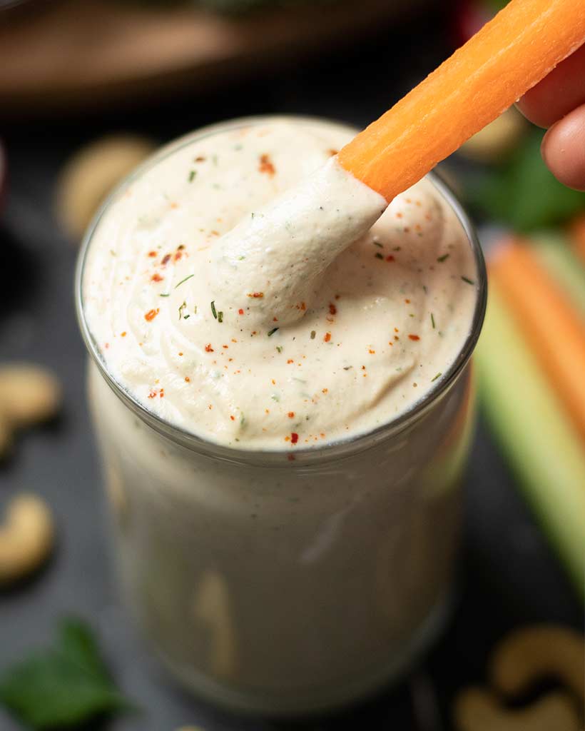 Carrot stick dipped in a creamy plant-based ranch dressing on table with raw cashews and freshly chopped veggies.