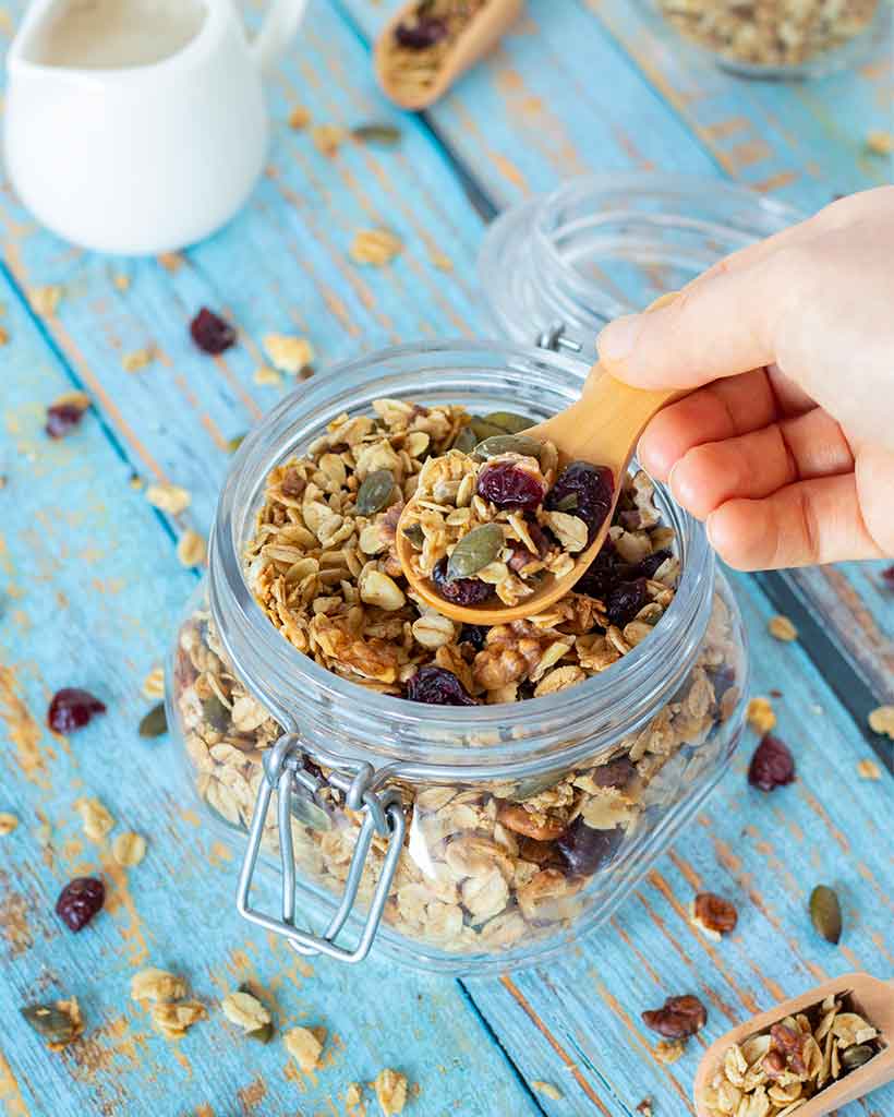 Homemade granola in a glass jar with wooden spoon on blue wooden table.