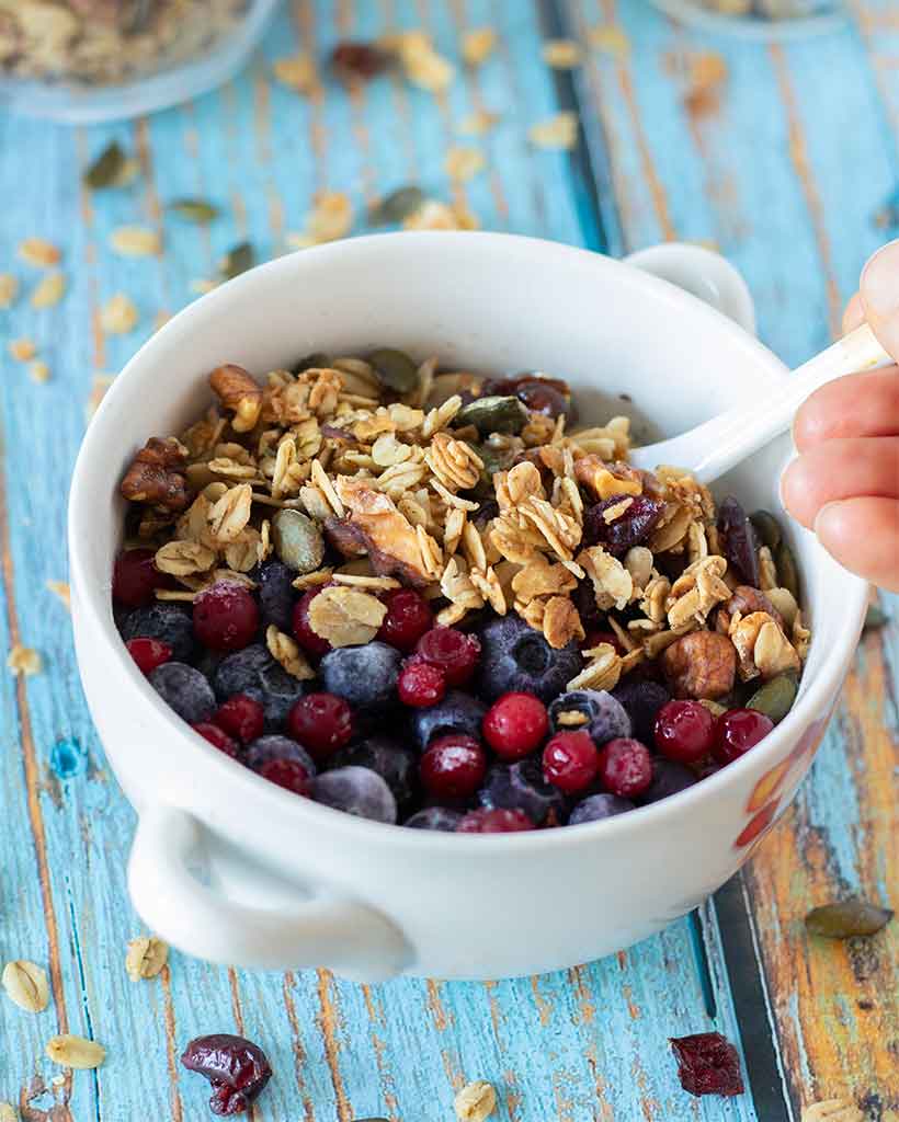 Healthy vegan granola breakfast dish with frozen blueberries and cranberries and dairy-free milk on blue wooden table.