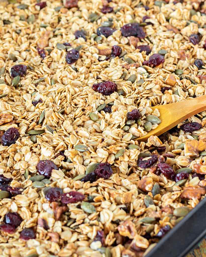 Homemade granola recipe healthy breakfast, snack, brunch or dessert idea. Gluten free freshly baked granola on a baking tray lined with parchment paper and a wooden spoon.