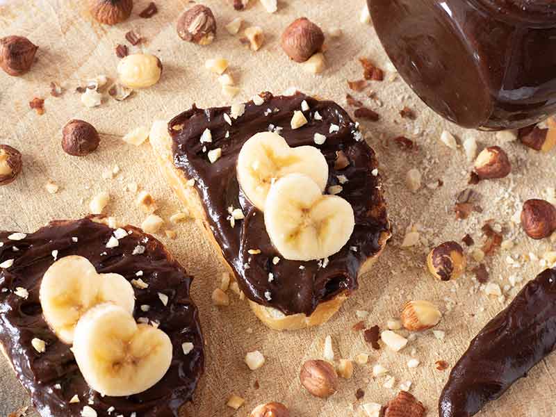 Healthy homemade Nutella with dates (no sugar, no palm oil, no milk) on a slices of bread with banana hearts and chopped hazelnuts.