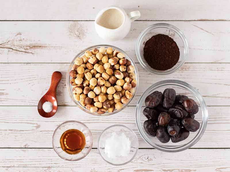 Simple, wholesome, plant-based ingredients for homemade Nutella recipe from scratch: roasted hazelnuts, dates, maple syrup, cacao powder, dairy-free milk, coconut oil, and salt.