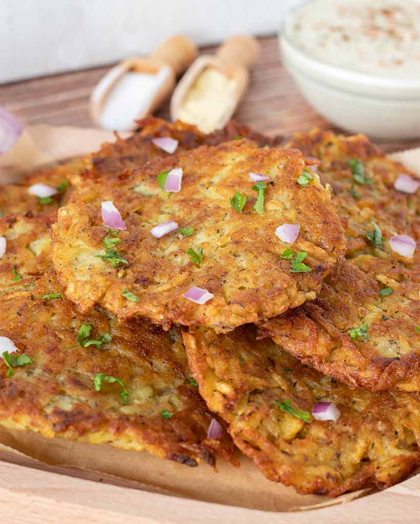 Golden-brown simple potato latkes (pancakes) on a wooden board decorated with chopped red onion, fresh parsley, and veggies as a side dish, breakfast, lunch or dinner.