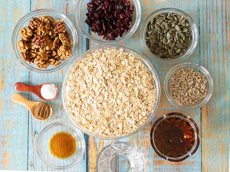 Simple, plant-based, budget-friendly ingredients for making homemade granola recipe: rolled oats, pepitas, walnuts, sunflower seeds, maple syrup, coconut oil, dried cranberries, vanilla extract, salt and ground cinnamon.