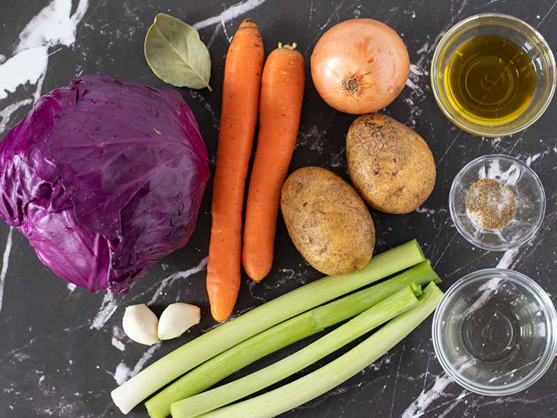 Simple, plant-based ingredients for cooking red cabbage soup recipe: fresh purple cabbage, potatoes, carrots, onion, leeks, garlic, olive oil, apple cider vinegar, and seasonings.