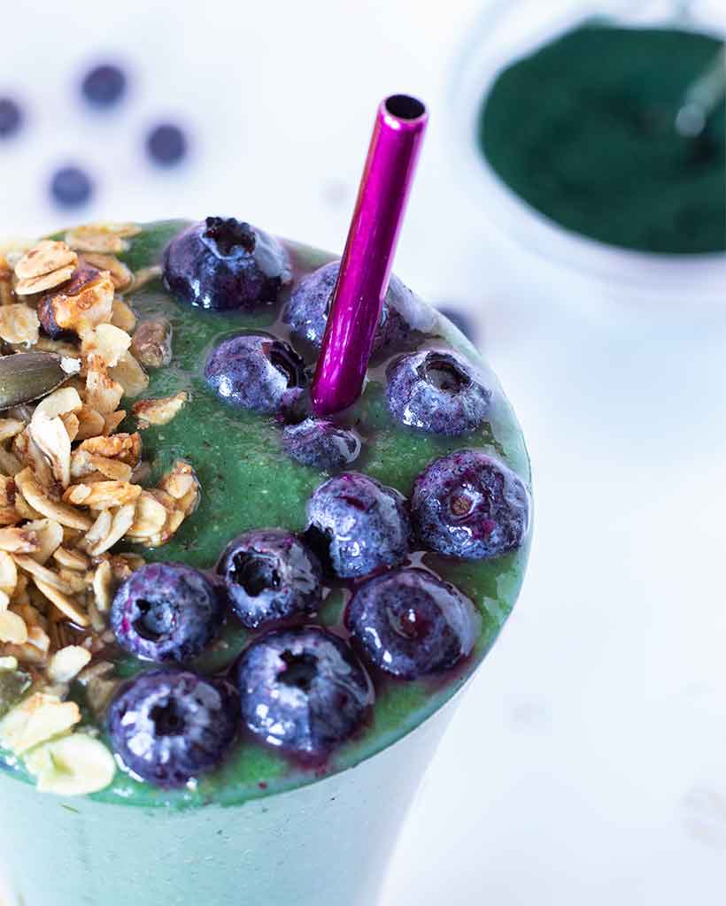 Green spirulina smoothie for weight loss and fat burning. Healthy and tasty morning breakfast drink. High-protein mix topped with frozen blueberries and homemade granola.