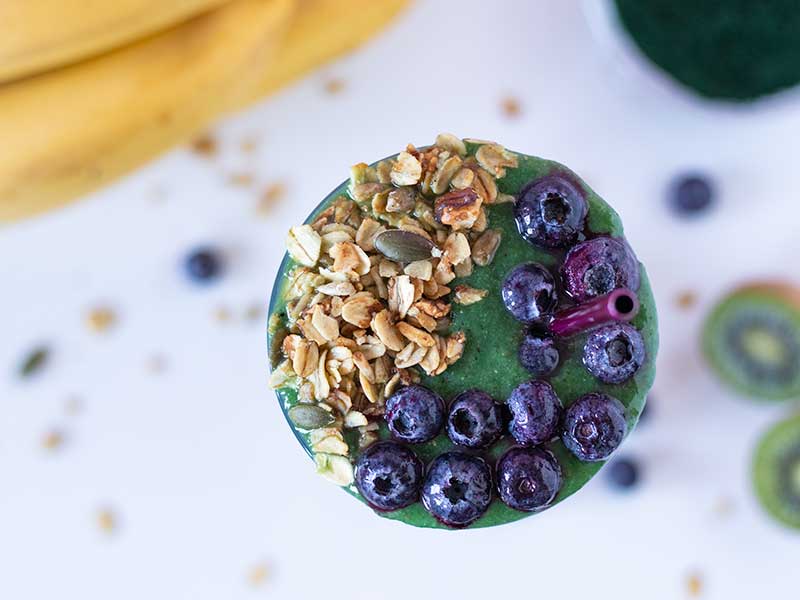 Bright green spirulina mix in a glass topped with blueberries and homemade granola.