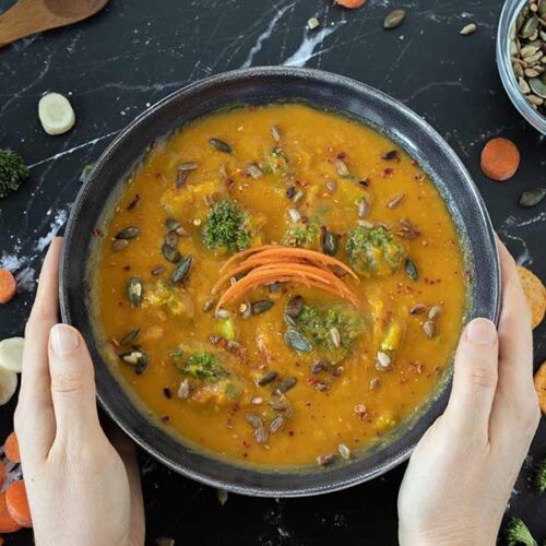 Easy vegan carrot and parsnip soup on dark granite table with woman's hands holding the dish and freshly chopped vegetables.