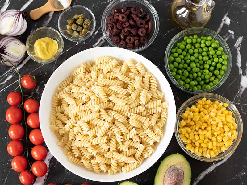 Simple plant-based ingredients for preparing tasty vegetarian pasta salad recipe: pasta, avocado, green peas, corn, olives, cherry tomatoes, olive oil, capers, mustard and easy dressing.