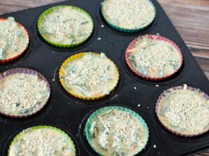 Muffin pan lined with silicone muffin liners filled with plant-based batter topped with sesame seeds.