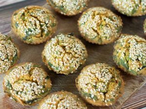 Baked savoury muffins topped with sesame seeds on wooden cutting board filled with healthy vegetables.