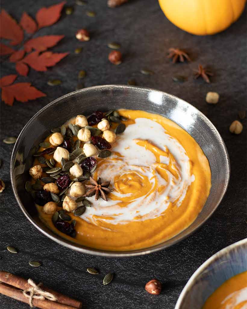 Colorful, immunity-boosting pumpkin puree soup on a dark table with red fall leaves, roasted hazelnuts, pumpkin seeds, cinnamon sticks, and star anise.