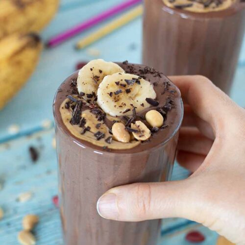 Creamy high-protein chocolate banana peanut butter smoothie in a glass with woman's hand holding it.