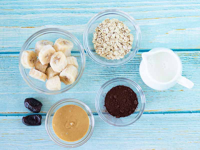 Simple plant-based ingredients for preparing easy and healthy smoothie blend: creamy peanut butter, dair-free milk, raw cacao powder, old-fashioned oats, frozen banana, and dates.