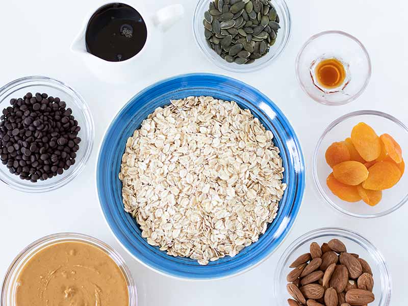 Simple, plant-based ingredients for preparing chewy homemade granola bars: rolled oats, peanut butters, vegan chocolate chips. almonds, dried apricots, pepitas, maple syrup and vanilla extract. 