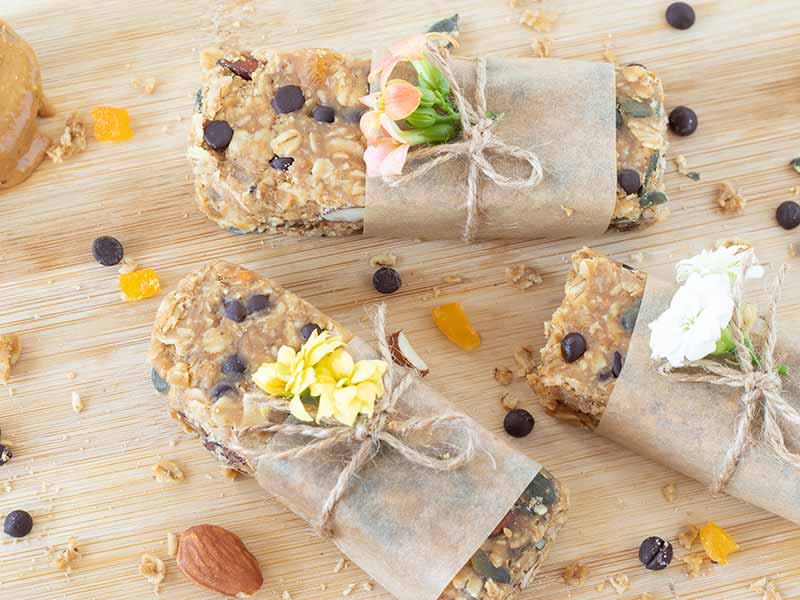 Easy DIY no bake-granola bars wrapped in parchment paper with small colorful flowers on wooden board for post-workout energy snack.