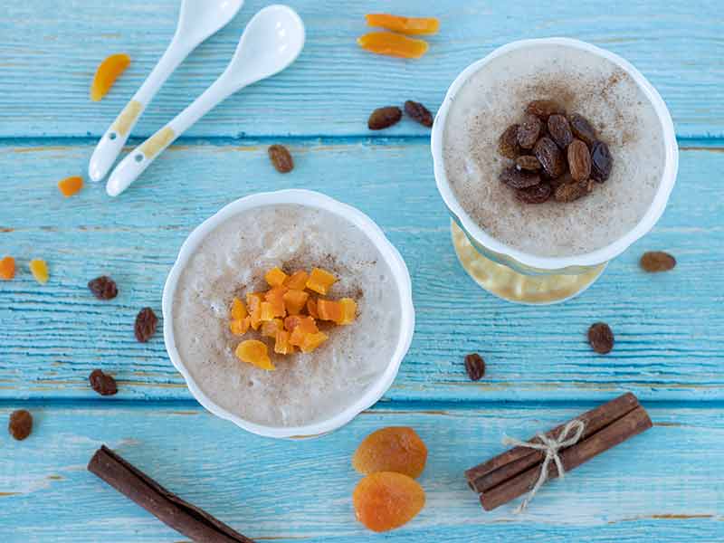 Egg-free creamy vegan rice pudding in small containers topped with ground cinnamon, dried apricots, and raisins on light blue wood table.