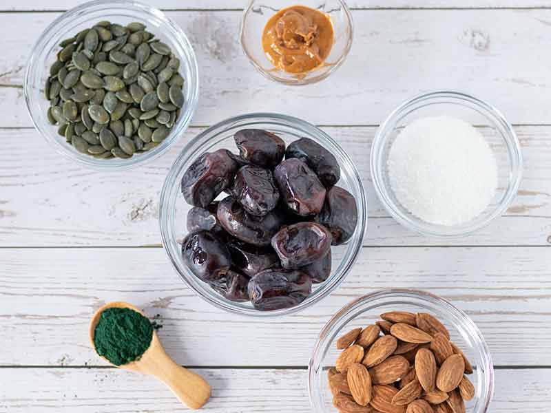 Sweet vegan energy balls in a plate on wooden table with spirulina powder, raw almonds and pepitas. Kid-friendly, dairy-free make ahead recipe without refined sugar or oil.