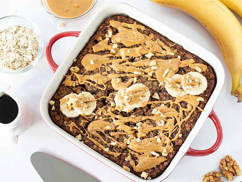 Baked oatmeal dessert cake in a baking pan drizzled with creamy peanut butter and topped with sliced bananas for an easy make-ahead breakfast meal prep at home.