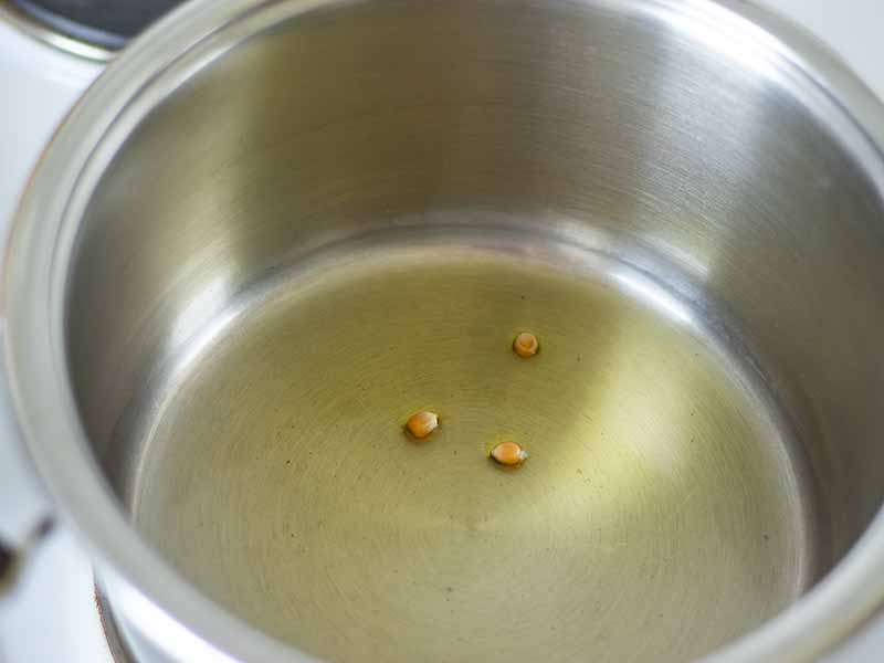 A large, stainless steel pot with olive oil and 3 corn kernels at the bottom prepared for making healthy homemade popcorn.