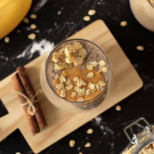 Oatmeal smoothie recipe, healthy vegan breakfast drink with banana and penut butter in a glass on a wooden cutting board with cinnamon sticks and rolled oats
