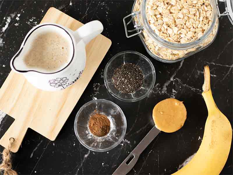 Wholesome plant-based ingredients for making tasty oat smoothie mix: rolled oats, banana, penut butter, dairy-free milk, chia seeds, ground cinnamon, and carob powder.