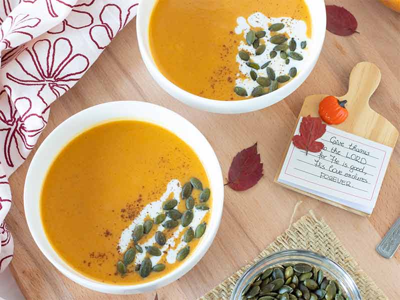 Best homemade vegan butternut squash soup in two bowls with handwritten note about thanksgiving to the LORD, autumn leaves in red color, and toasted pumpkin seeds on a wooden table.