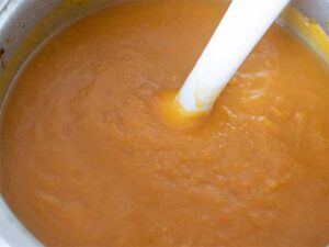 An immersion blender in a pot with hot autumn soup with orange color
