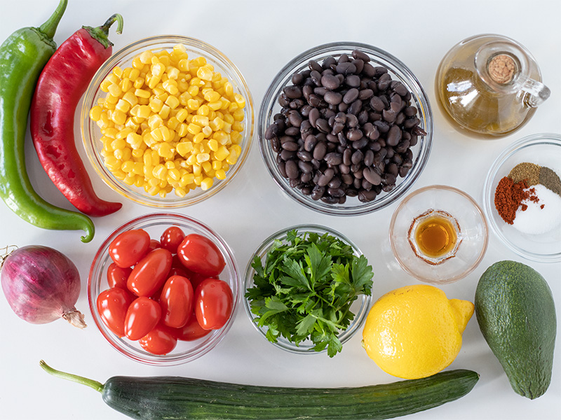 Fresh plant-based ingredients for preparing easy vegan salad: black beans, corn, avocado, plum cherry tomatoes, parsley, red and green peppers, cucumber, red onion, extra virgin olive oil, pure maple syrup, freshly squeezed lemon juice, and spices