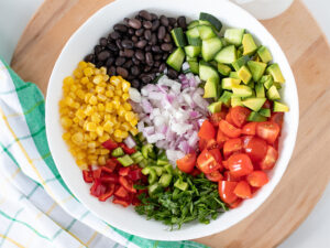 Finely chopped fresh plant-based ingredeints in salad bowl: corn kernels, black beans, green and red pepper, cucumber, avocado, red onion, cherry tomatoes, and parsley.
