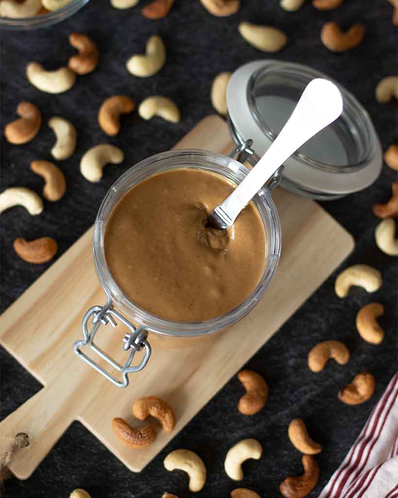 Easy DIY recipe for cashew butter. Creamy nut spread in a jar with spreading knife on dark background with raw and roasted cashews for high-protein breakfast or snack.