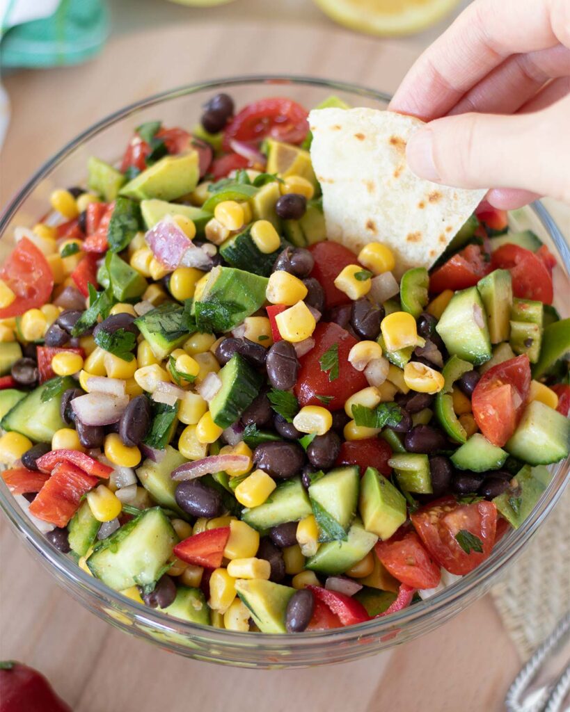 Female hand holding homemade torilla dipped in a big bowl of fresh  corn salad with black beans and tomato