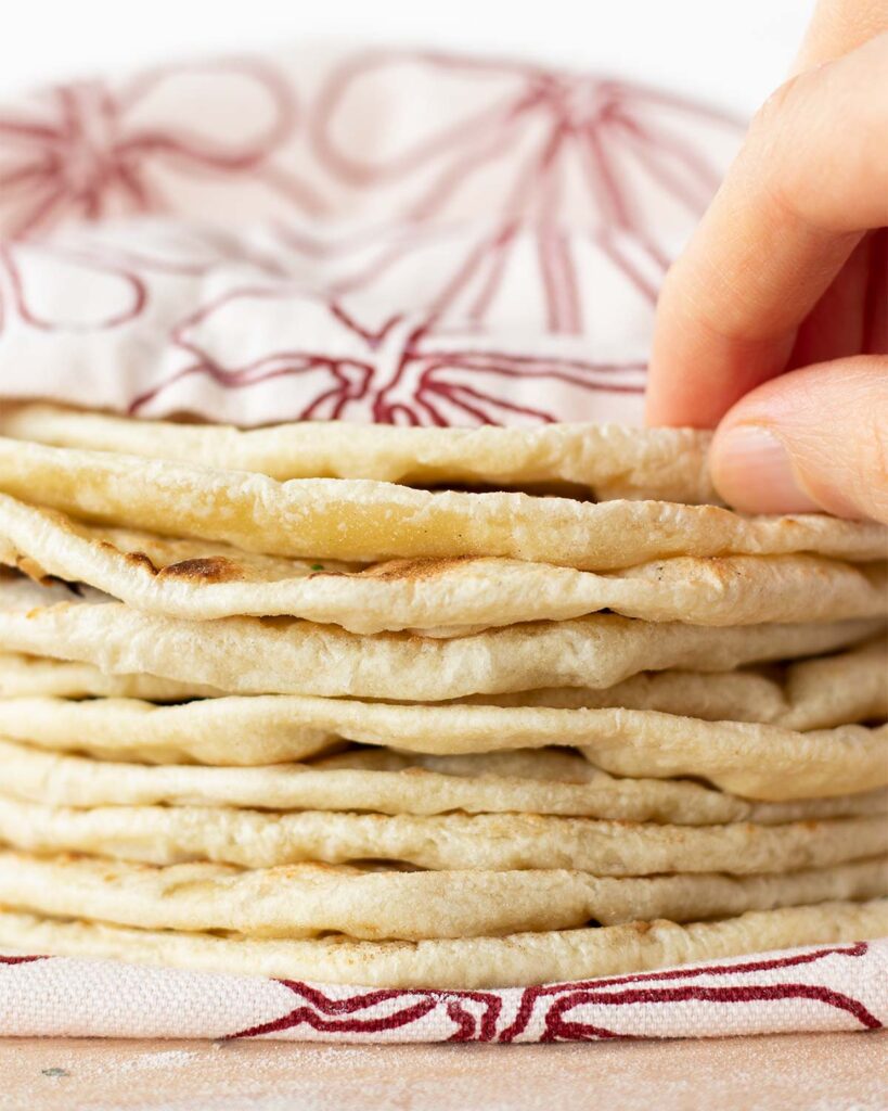 Stacked homemade flour tortillas covered with kitchen towel on wooden table