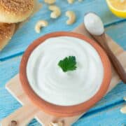 Best vegan sour cream in a rustic bowl decorated with persley leaf on wooden cutting board with raw cashews, spoon, and homemade bagels