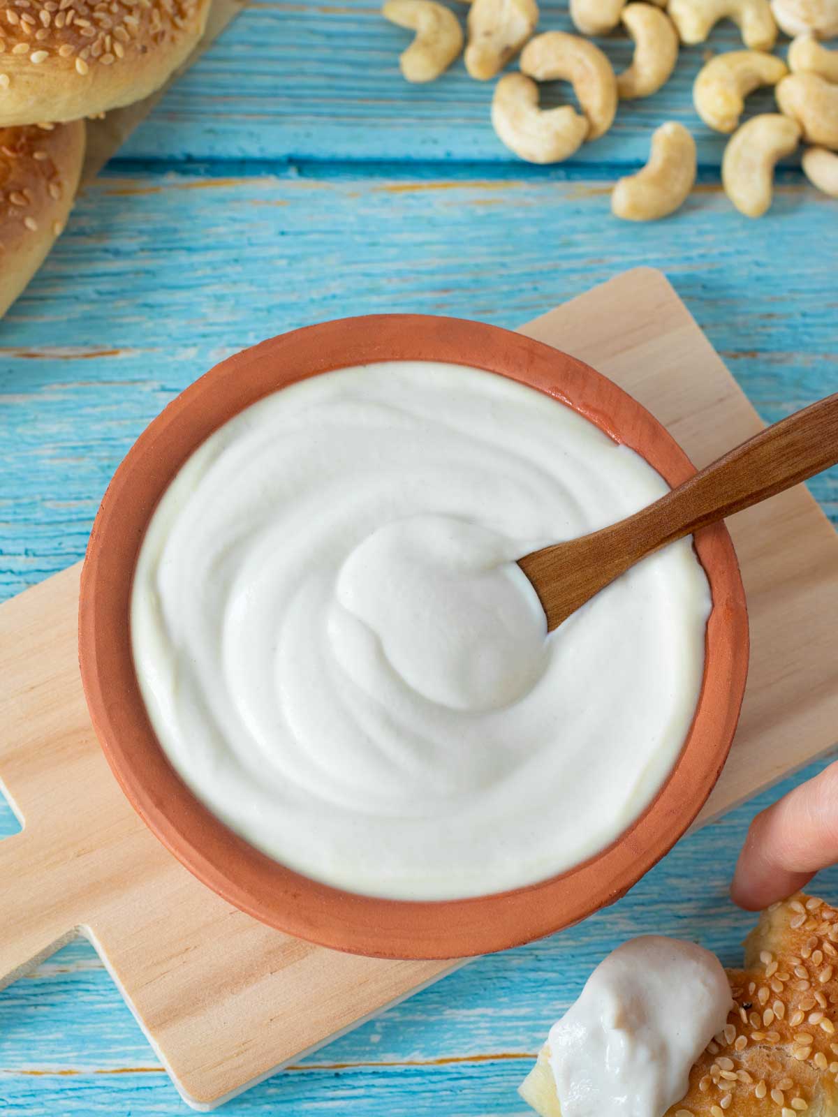 Homemade dairy-free sour cream in a bowl with wooden spoon, raw cashews, and bagel on wooden table