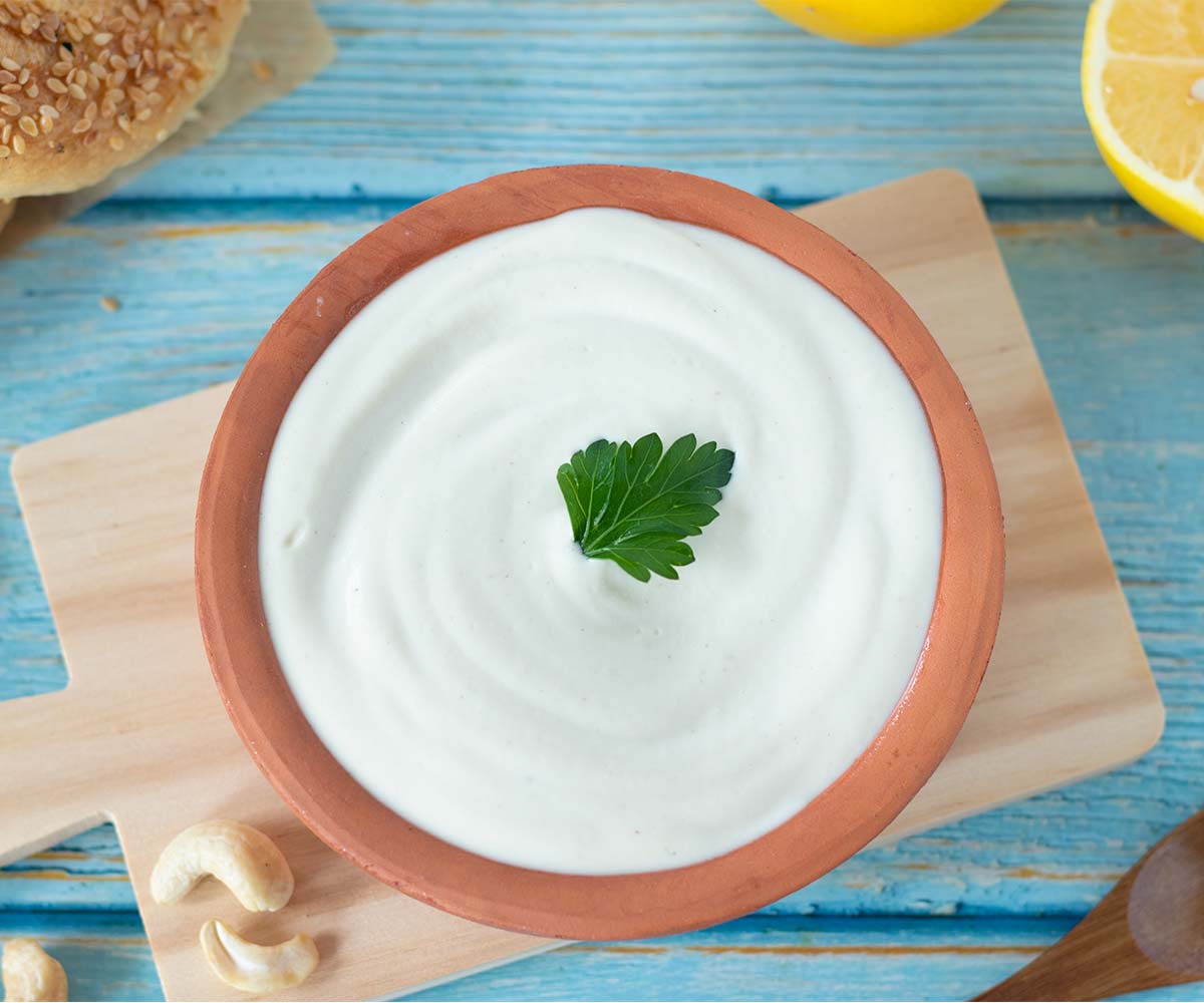 Creamy non-dairy sour cream made with cashews in a bowl decorated with fresh parsley leaf, a wooden spoon, lemon and bagen on table