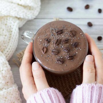 Woman's hands holding creamy chocolate coffee smoothie in a cup on table with woolen scarf