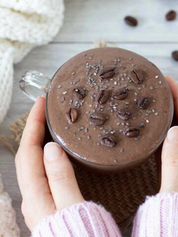 Woman's hands holding creamy chocolate coffee smoothie in a cup on table with woolen scarf