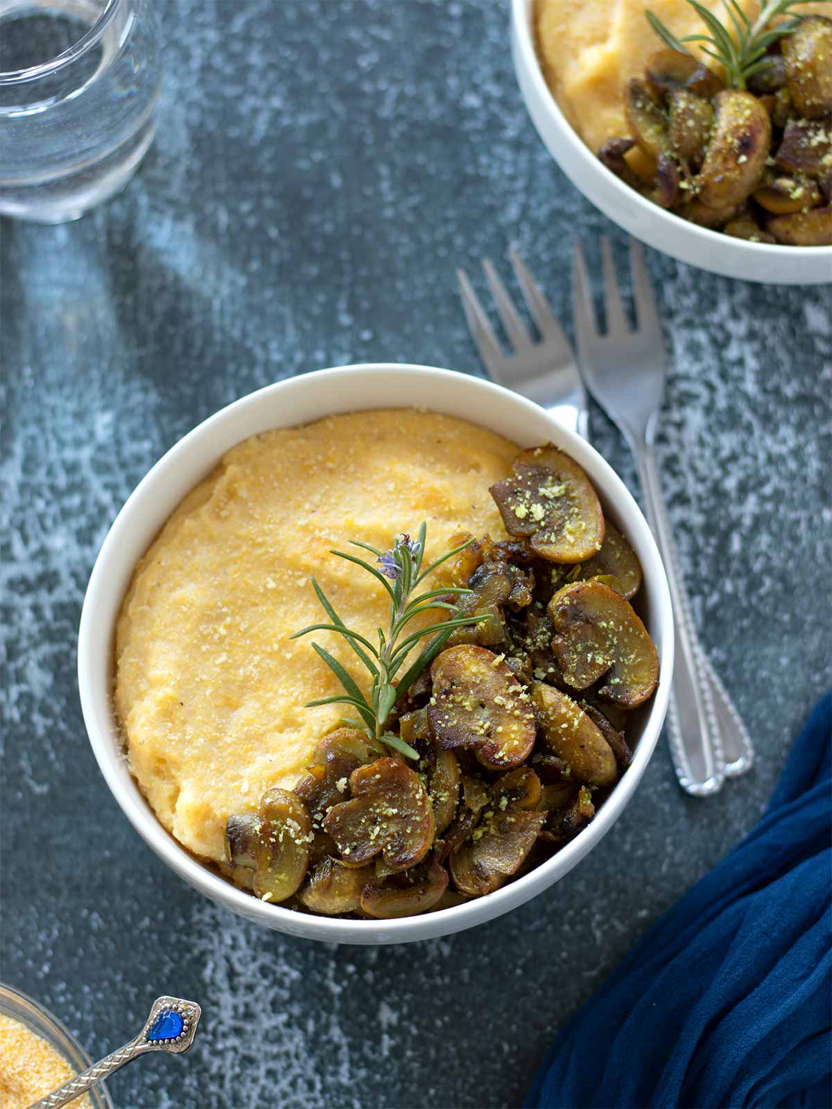 Creamy mushroom polenta in a bowl garnished with fresh rosemary with forks and a glass of water