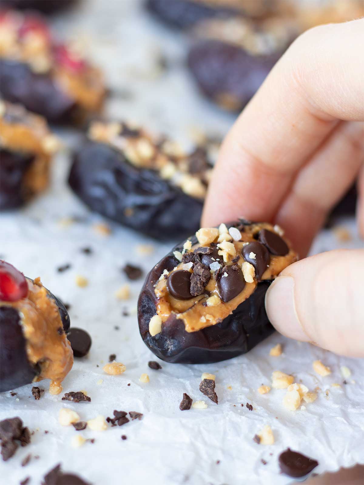 A close-up of female hand holding a peanut butter stuffed date topped with mini chocolate chips, chopped dark chocolate and chopped roasted peanuts