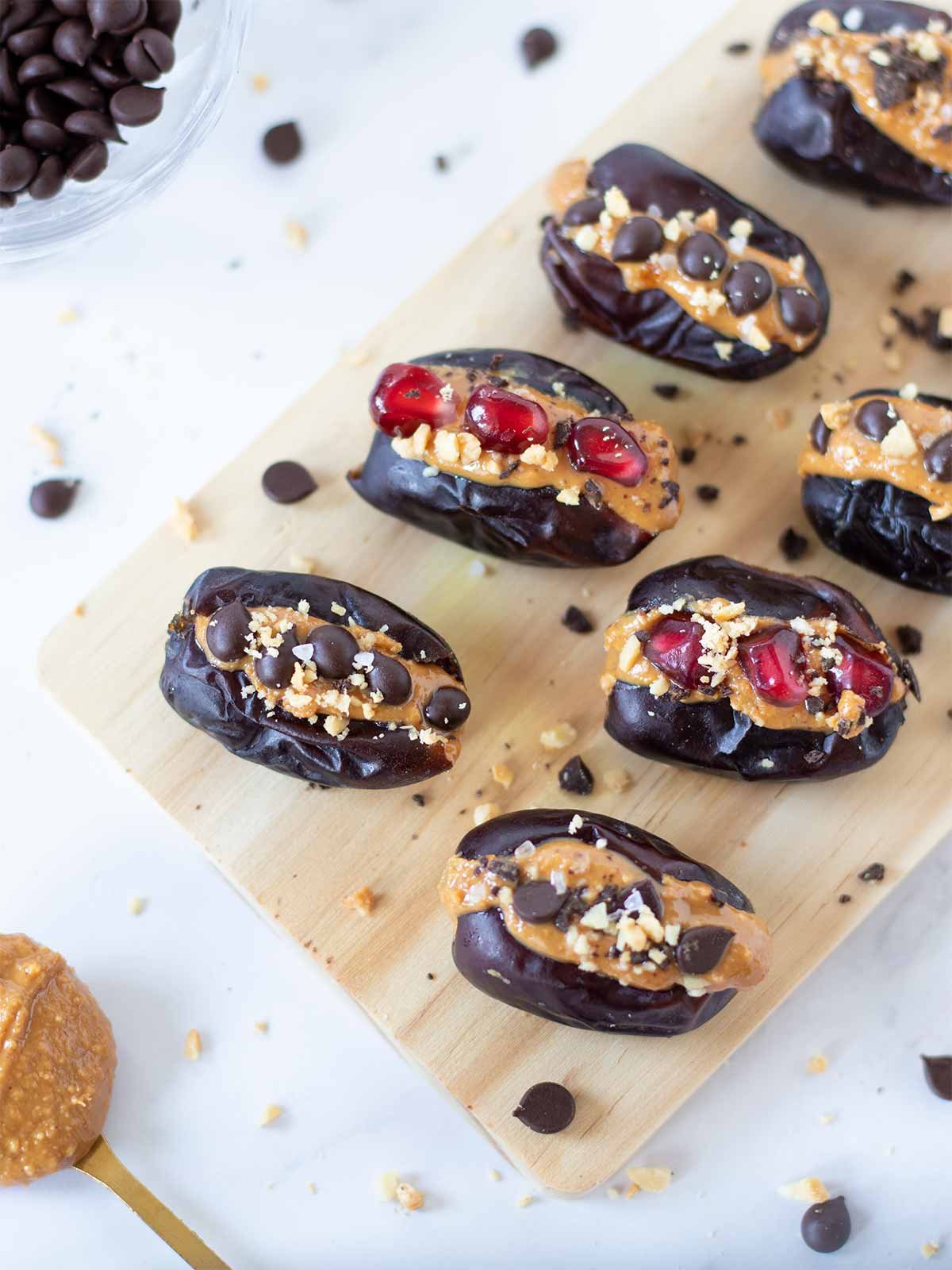 Mazafati dates filled with natural creamy peanut butter decorated with mini chocolate chips, chipped dark chocolate, chopped peanuts, and pomegranate arils on wooden board